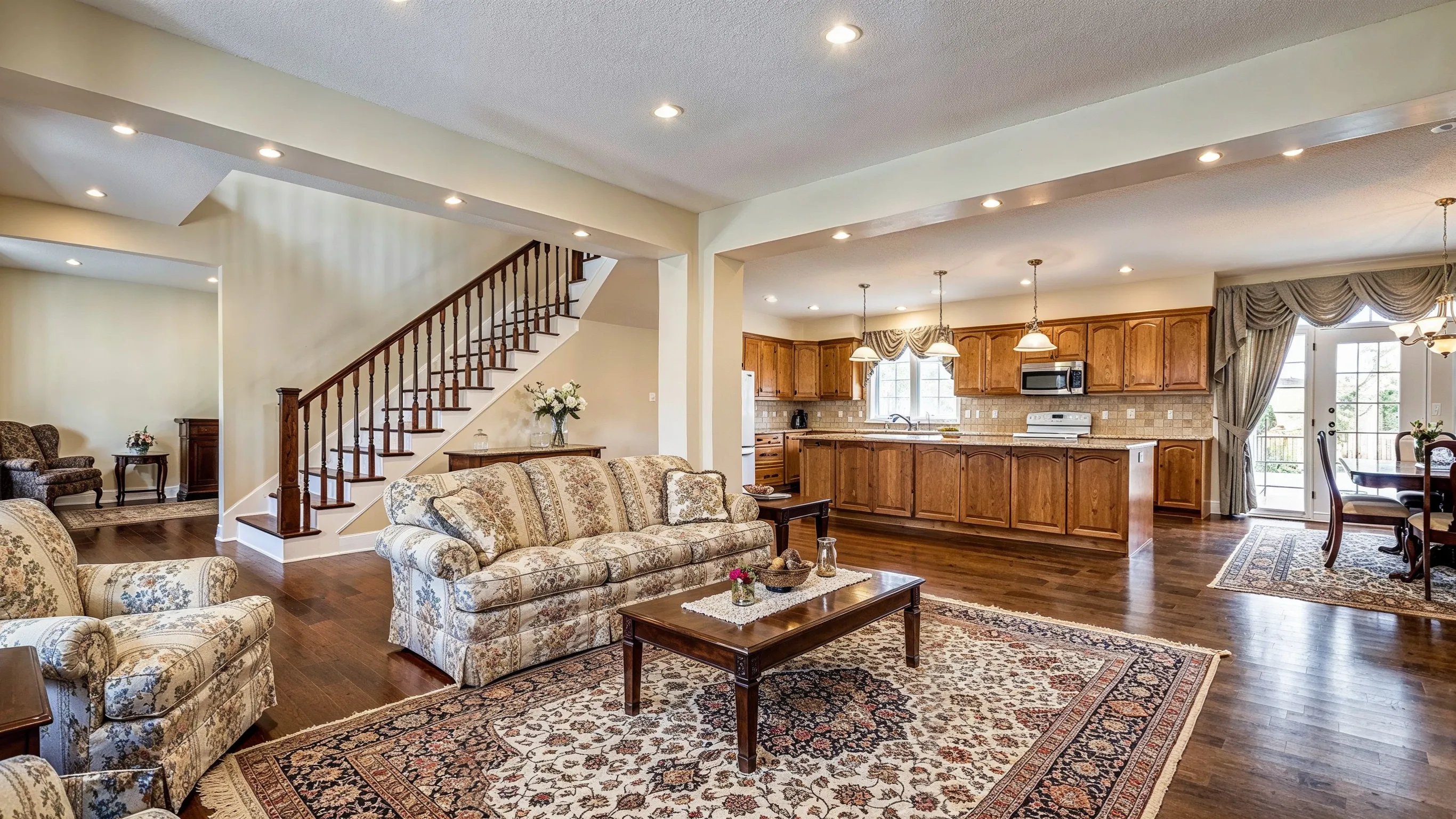 Before photo of luxury home remodeling project in Los Angeles with traditional living room, older wood kitchen cabinets, classic staircase, patterned furniture, dark flooring, and dated interior finishes
